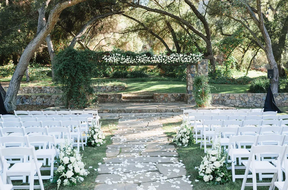 Rows of white chairs on a lawn under the spreading canopy of mature oak trees, a petal-strewn aisle down the center.