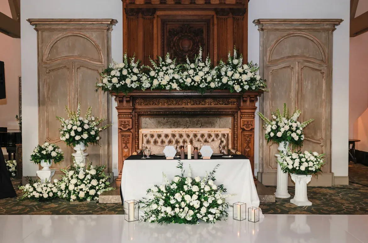 A sweetheart table set before a stone fireplace banked with white florals and low candlelight.