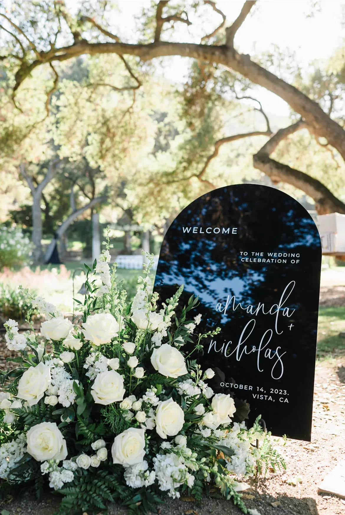 A hand-lettered welcome sign in white script on matte charcoal, set in a low spray of white garden roses.