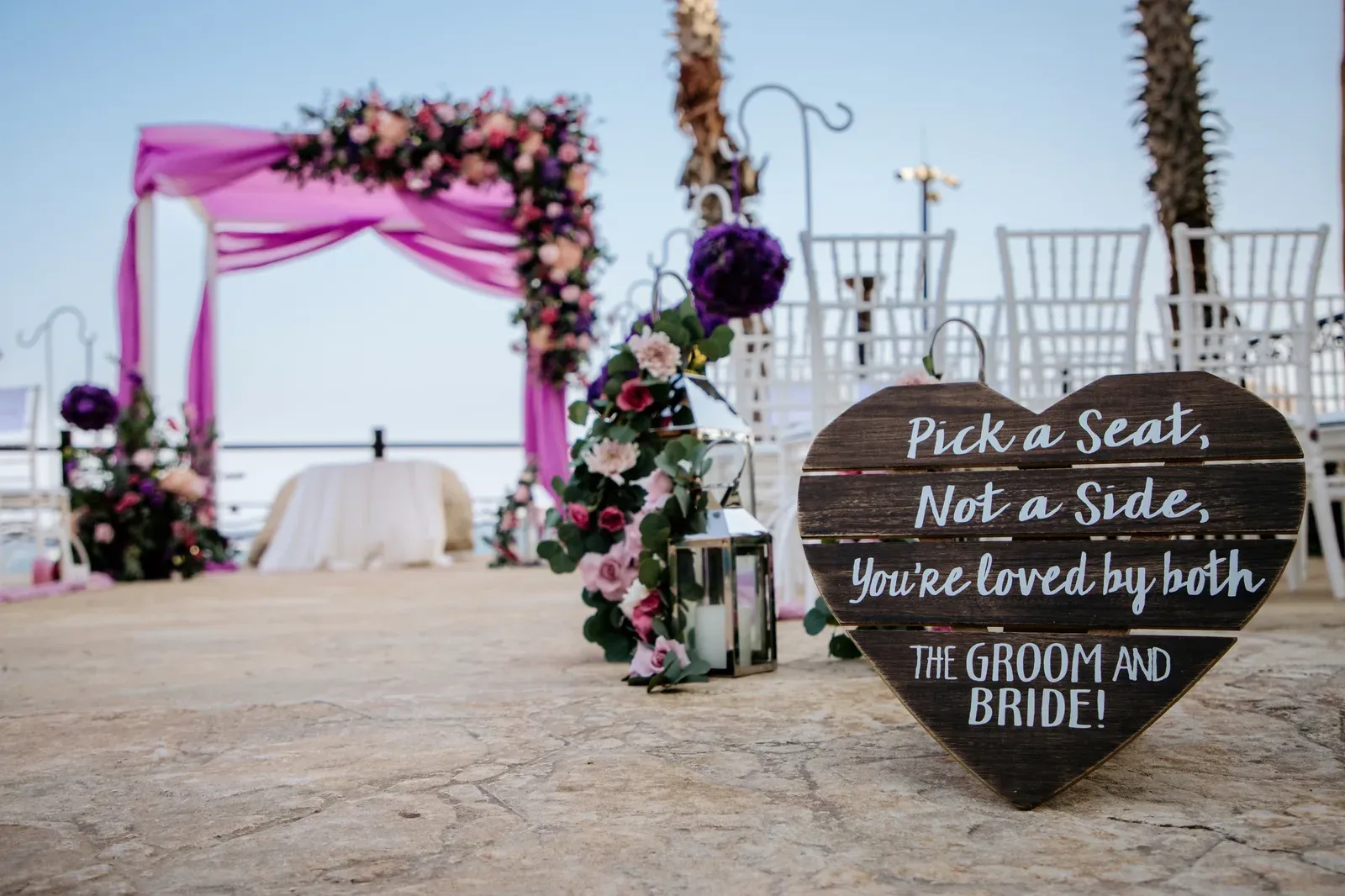 A ceremony aisle lined with white chairs and purple floral pomanders, with a draped arch at the water.