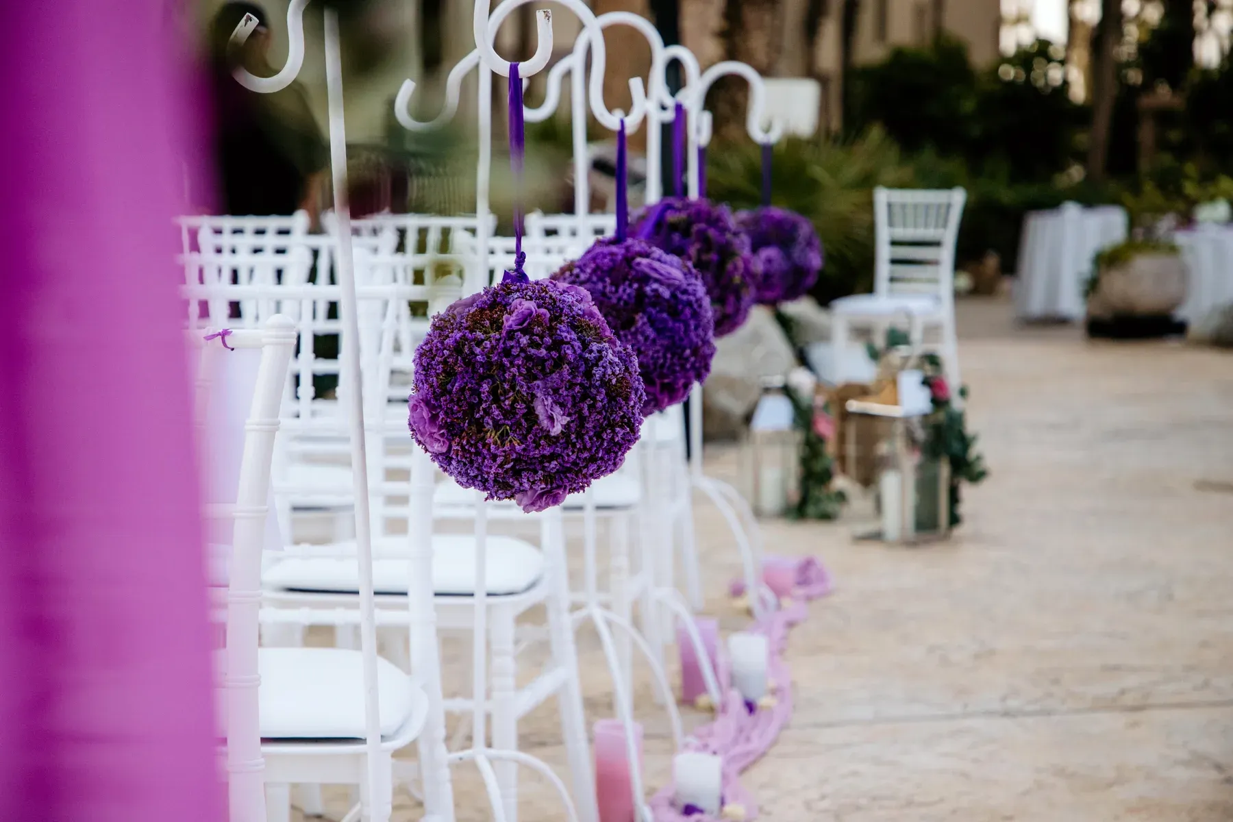 Purple floral pomanders hanging from white chairs along a petal-strewn oceanfront ceremony aisle.
