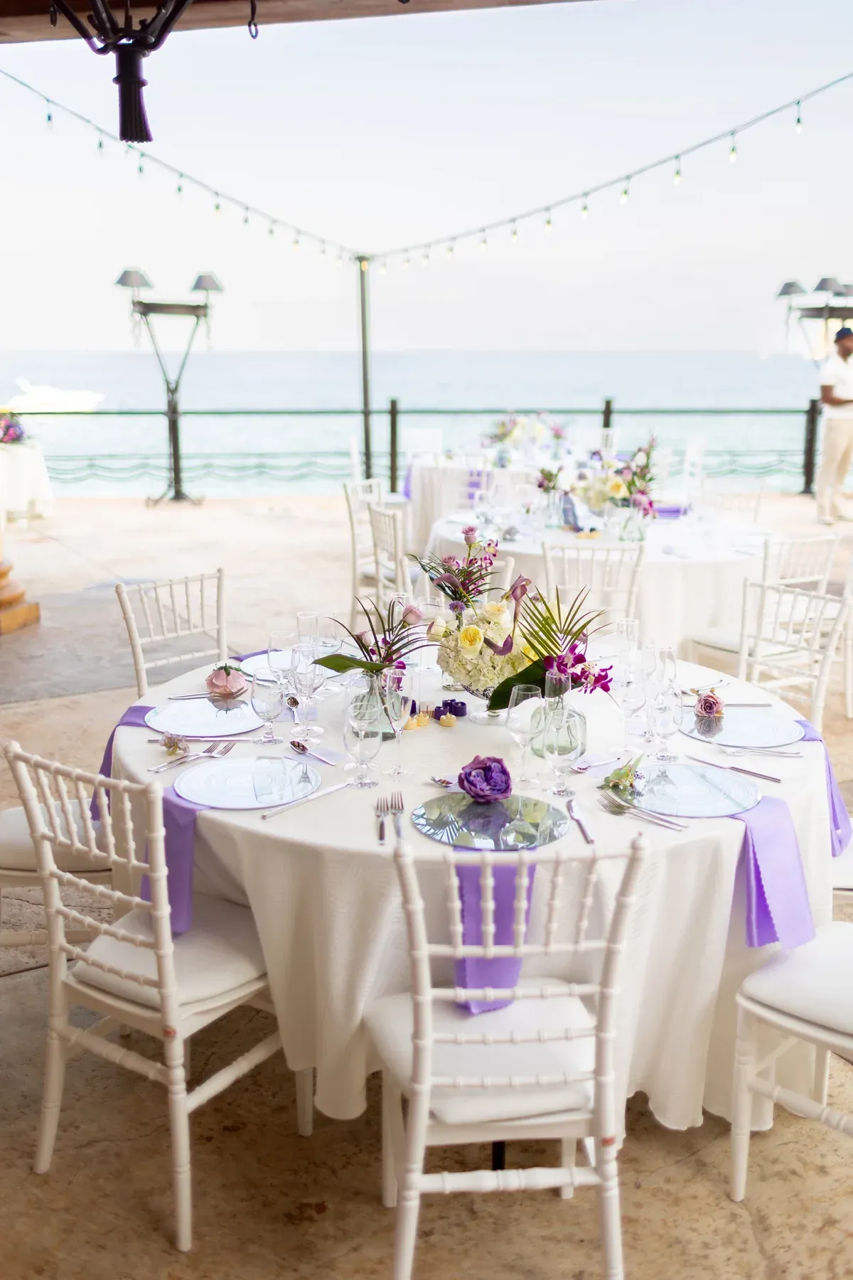 A round guest table set with pale blue glass chargers and low purple florals, the ocean behind.