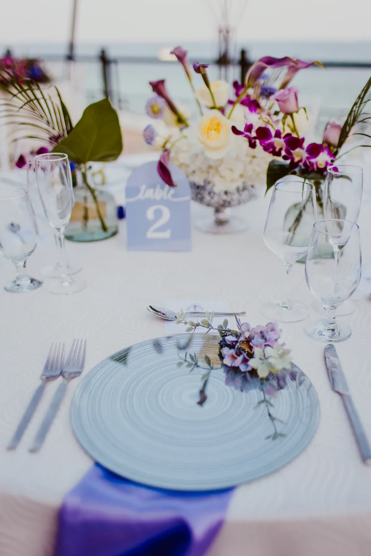 A place setting with a pale blue glass charger, a sprig of orchid, and a slate-blue table number.